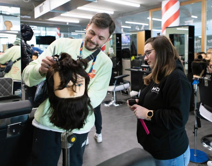 A male instructor in a light green sweatshirt assisting a female student with a mannequin head in a barbering classroom. A male instructor in a light green sweatshirt assisting a female student with a mannequin head in a barbering classroom.